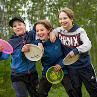 Kolme nauravaa nuorta frisbeet kädessä, kädet toistensa hartioilla