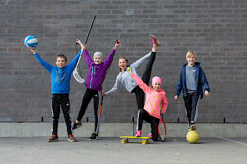 Lapsilla eri liikunnan harrastusvälineitä käsissä esim. koripallo, skeitti, tennismaila ja tuulettavat iloisesti