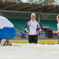 Hymyilevä valmentaja katsoo ja tsempaa nuoria urheilijoita korkeushyppypaikalla