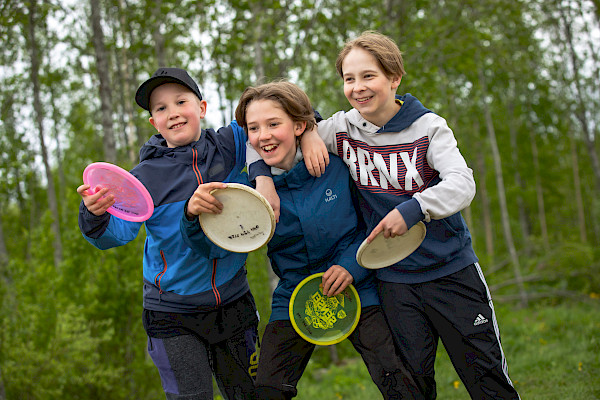 Kolme nauravaa nuorta frisbeet kädessä, kädet toistensa hartioilla