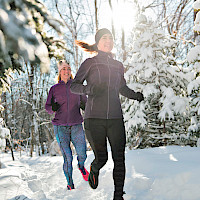 Kaksi henkilöä juoksee lumisessa metsässä auringonpaisteessa