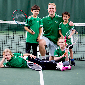 Tennisvalmentaja ja lapsi valmmenettavat hymyilevät kameralle tennisverkon edessä