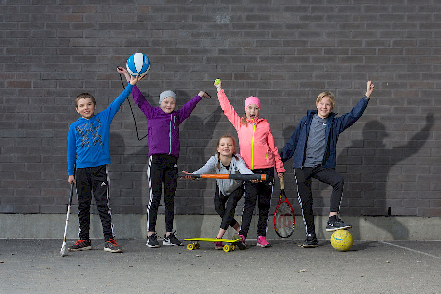 Hymyileviä koululaisia eri liikuntavälineet kädessä: pesäpallomaila, koripallo, skeitti, hyppynaru