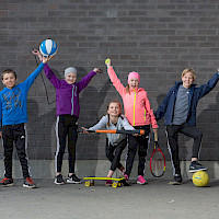 Hymyileviä koululaisia eri liikuntavälineet kädessä: pesäpallomaila, koripallo, skeitti, hyppynaru